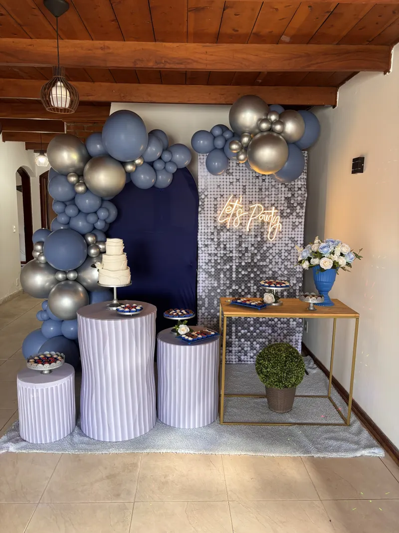 Mesa do bolo com arco de balões azuis e prateados, painel azul-marinho e painel de paetês com neon “Let’s Party”
