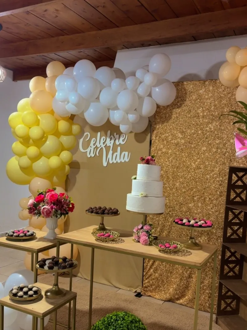Mesa do bolo com balões amarelos, brancos e cor de pêssego, painel dourado e o letreiro “Celebre a Vida”